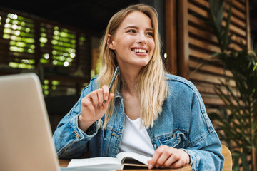 Happy young woman student posing outdoors in cafe using laptop computer writing notes in notebook.