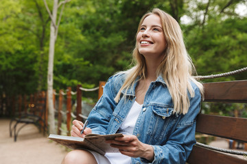 Amazing happy young woman student posing outdoors in park writing notes in notebook.