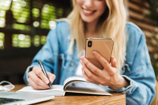 Happy young woman student posing outdoors in cafe using laptop computer writing notes in notebook.
