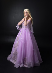 Fototapeta premium full length portrait of a blonde girl wearing a fantasy fairy inspired costume, long purple ball gown with fairy wings, standing pose on a dark studio background.