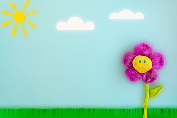 Paper sun, clouds, green grass and toy big flower with a smiling face on a blue background. Funny summer landscape composition on the theme of good sunny weather and good mood.