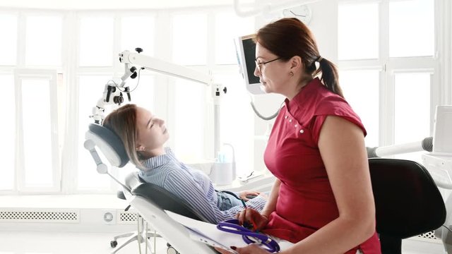 A young girl at a consultation with a dentist woman in glasses is sitting on a chair in a stamotology office. The conversation of the doctor and the patient