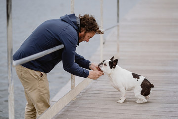 Side view of adult man in casual outfit petting adorable French Bulldog while spending time on lumber pier near sea
