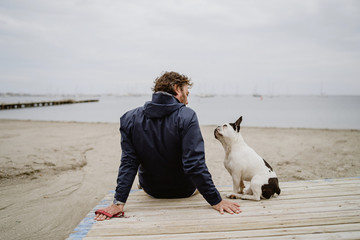 Adult male in warm jacket embracing spotted French Bulldog while sitting on wooden pier and admiring view of rippling sea on dull day