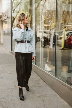 Attractive young female in stylish outfit doing hair and looking at clothes behind shop window while walking on city street