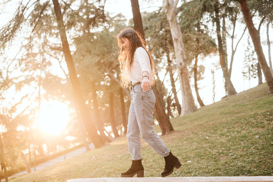 Side view of young female in casual outfit stretching out arms and trying to keep balance while walking on border on sunny day in park