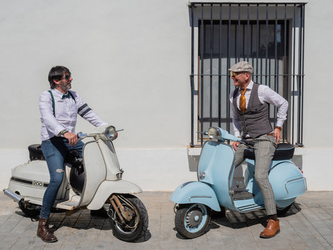 Positive middle-aged hipsters in elegant clothes with retro motorbikes looking at each other in sunny day