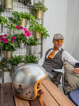 Pensive middle-aged hipster in elegant clothes sitting at wooden table with motorcycle helmet using smartphone