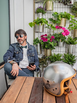 Pensive middle-aged hipster in elegant clothes and sunglasses sitting at wooden table with motorcycle helmet using smartphone