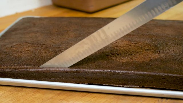 Baker Cuts Off The Edge Of A Chocolate Cake In Pro Kitchen. Professional baker uses a large knife to cut the edges of a chocolate cakes to make it even and square.