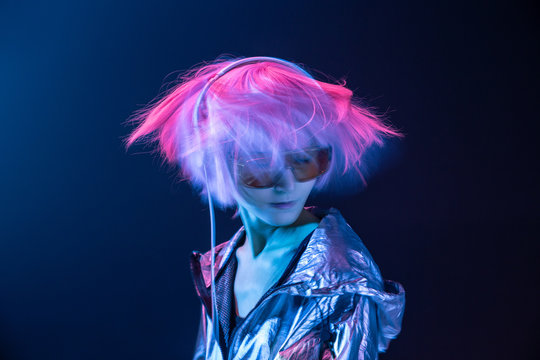 Young subcultural Asian woman with pink scattered hair and silver jacket dancing while listening to music in headphones