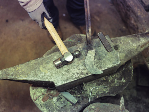 Crop blacksmith holding metal with equipment on anvil with hammer in traditional workshop