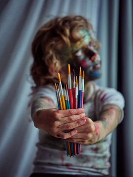 Pretty creative boy with blond hair painted in colorful paints holding paintbrushes on outstretched hands and looking away