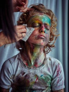 Crop young beautiful female with napkins cleaning pretty creative boy with blond hair smeared in colorful paints