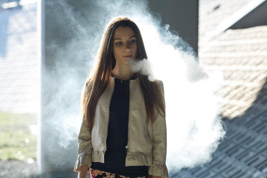 Vape teenager. Young cute girl in  casual clothes smokes an electronic cigarette outdoors in summer day. Bad habit that is harmful to health. Vaping activity.