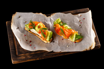 Italian bruschetta with salmon and cucumber at wooden board isolated at black background.