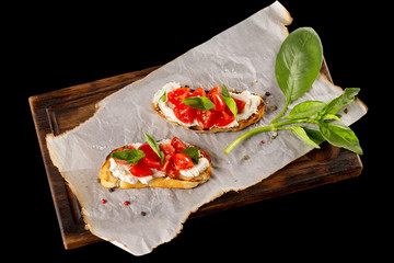 Italian bruschetta with cream cheese, tomatoes and basil at wooden board isolated at black background.