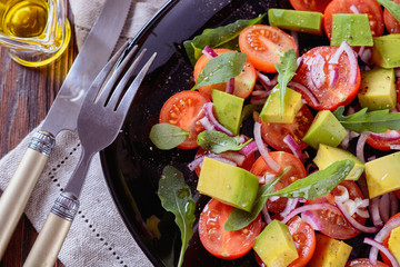 Colorful and tasty salad with avocado, cherry tomatoes and mozzarella on a black dish next to oil in glass sauce boat, fork and knife, top view