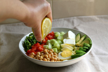 Squeezing lemon juice on lunch bowl with avocado, hard boiled egg, chickpea, feta cheese, cucumber, tomato and arugula. Selective focus.
