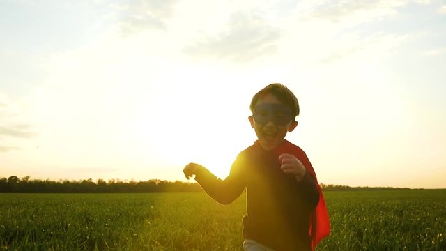 The child, dressed as a superhero in a red cloak, runs across the green grass against the sunset, rejoices and smiles