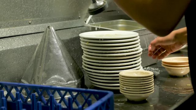 young man washes dishes in professional kitchen with sprayer. Employee washes and rinses the front and back of a pile of dirty plates and places them in a drying rack. Employee uses a professional kit