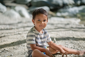 Cute 3 year old child wearing a striped shirt and shorts, sitting on the beach with the river in the background on a sunny day.