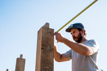 Man wearing baseball cap and sunglasses on building site, using tape measure.
