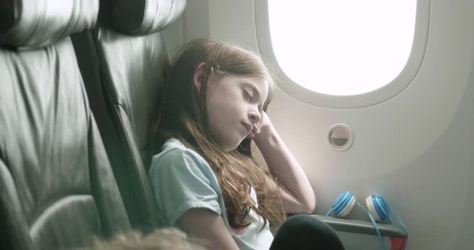young girl sleeps with her head resting against the bulkhead on a plane as the camera pans around her
