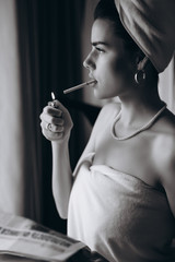 Beautiful young woman in a towel smokes a cigarette and reads newspaper