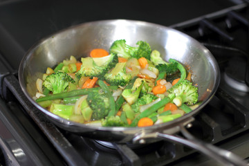 Stir fry vegetables cooking in a stainless steel pan on a natural gas stove.