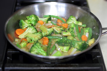 Stir fry vegetables cooking in a stainless steel pan on a natural gas stove.