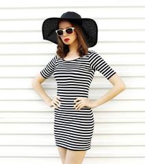 Stylish young woman in striped dress, summer straw hat posing on white wall background