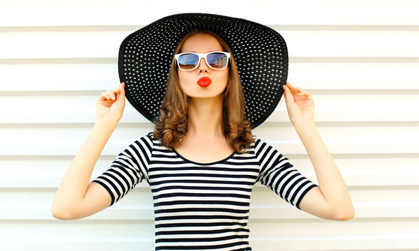 Portrait young woman blowing red lips sending sweet air kiss in black summer straw hat on white wall background