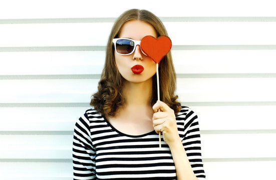 Portrait close-up woman blowing red lips sending sweet air kiss hiding her eye with red heart shaped lollipop on white wall background