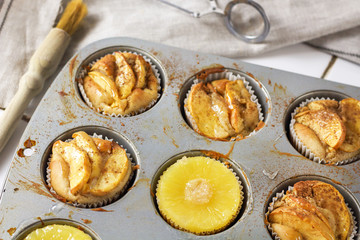 Gluten-free homemade vegetarian desserts cupcakes muffins with apple slices and pineapple rings on top in baking tray in kitchen on white-tiled worktop, next to a kitchen tea-strainer, towel