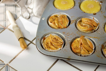 Gluten-free homemade vegetarian desserts cupcakes muffins with apple slices and pineapple rings on top in baking tray in kitchen on white-tiled worktop, next to a kitchen tea-strainer, towel
