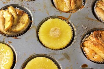 Gluten-free homemade vegetarian desserts cupcakes muffins with apple slices and pineapple rings on top in baking tray in kitchen on white-tiled worktop, next to a kitchen tea-strainer, towel