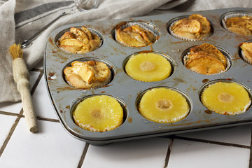 Gluten-free homemade vegetarian desserts cupcakes muffins with apple slices and pineapple rings on top in baking tray in kitchen on white-tiled worktop, next to a kitchen tea-strainer, towel