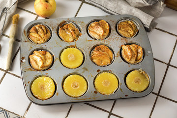 Gluten-free homemade vegetarian desserts cupcakes muffins with apple slices and pineapple rings on top in baking tray in kitchen on white-tiled worktop, next to a kitchen tea-strainer, towel