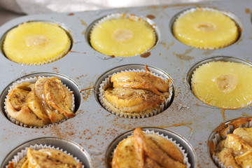 Gluten-free homemade vegetarian desserts cupcakes muffins with apple slices and pineapple rings on top in baking tray in kitchen on white-tiled worktop, next to a kitchen tea-strainer, towel
