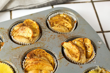 Gluten-free homemade vegetarian desserts cupcakes muffins with apple slices and pineapple rings on top in baking tray in kitchen on white-tiled worktop, next to a kitchen tea-strainer, towel