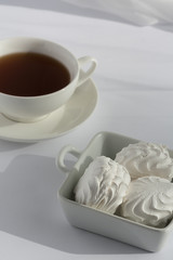 Square deep plate full of marshmallows and white cup of black tes on a plate on a white with veil on the background. Natural daylight.