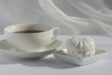 One marshmallow on a squre small plate and white cup of black tea on a plate on a white with veil on the background. Natural daylight.