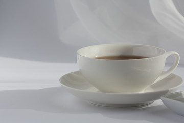 Cup of tea in white cup on a white plate. White veil in the background. Natural daylight, elegant close up morning scene.