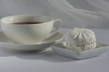 One marshmallow on a squre small plate and white cup of black tea on a plate on a white with veil on the background. Natural daylight.