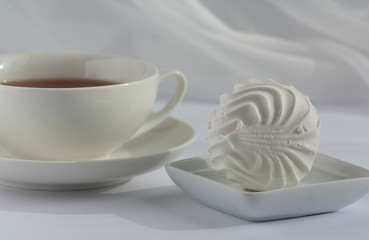 One marshmallow on a squre small plate and white cup of black tea on a plate on a white with veil on the background. Natural daylight.