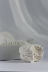 Obraz premium One bitten marshmallow with full square deep plate of marshmallows on white background. Natural daylight.