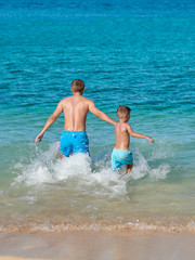 European boy and his dad are running in to the ocean holding hands, they are making a lot of splashes around.