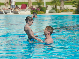 Handsome European man and his son are having fun in the swimming pool. They are enjoying their summer holidays.