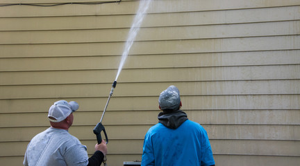 Two young men standing by the side of a yellow house. One is power washing the siding with a long...